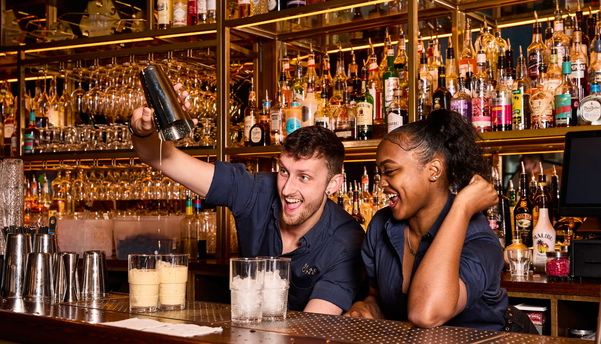 Bartenders mixing cocktails behind the bar at Be At One Liverpool Street with shelves of spirits