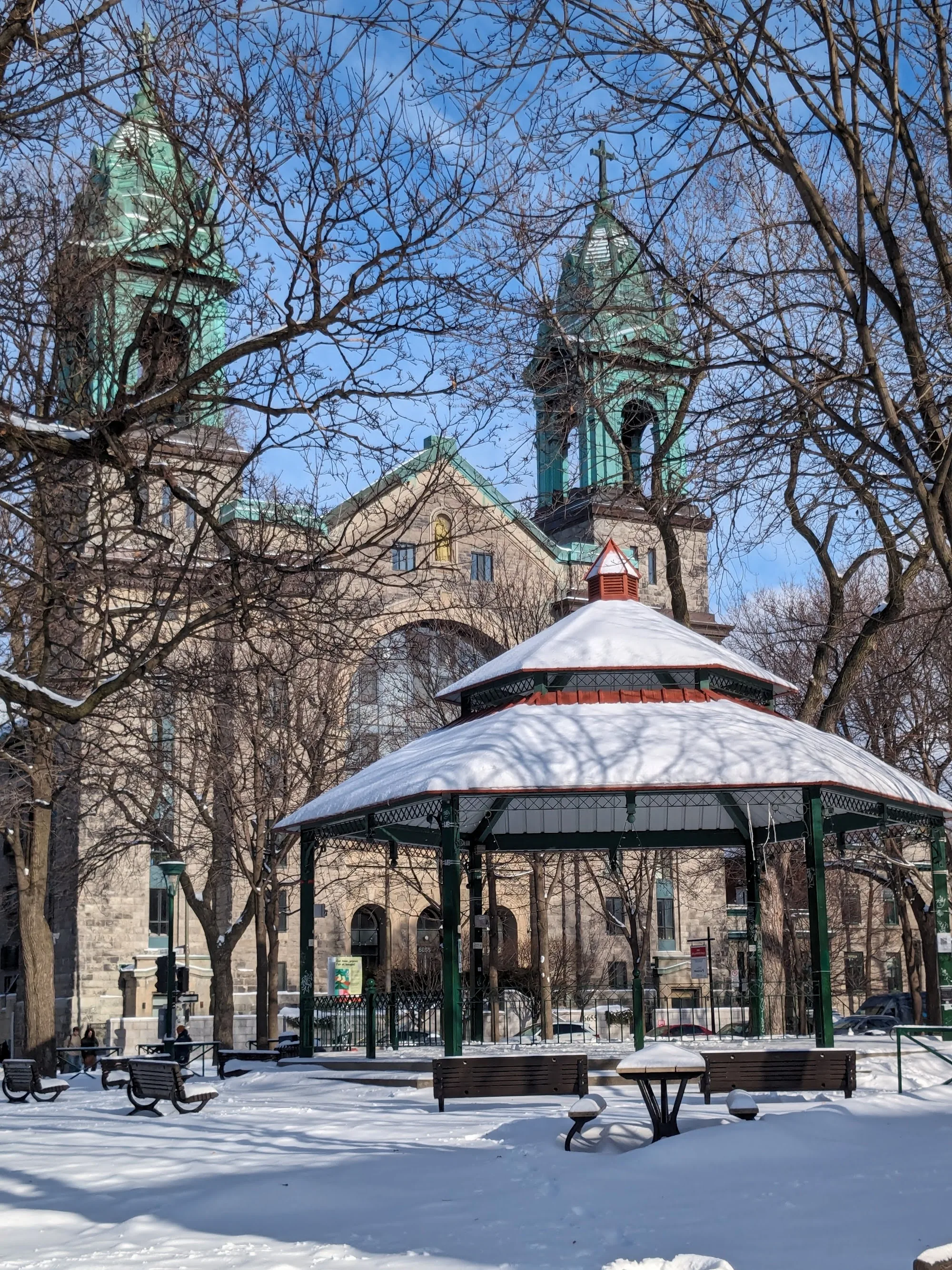Park of Little Italy Montreal in winter — gazebo bandstand with Église Saint-Zotique church towers and snow