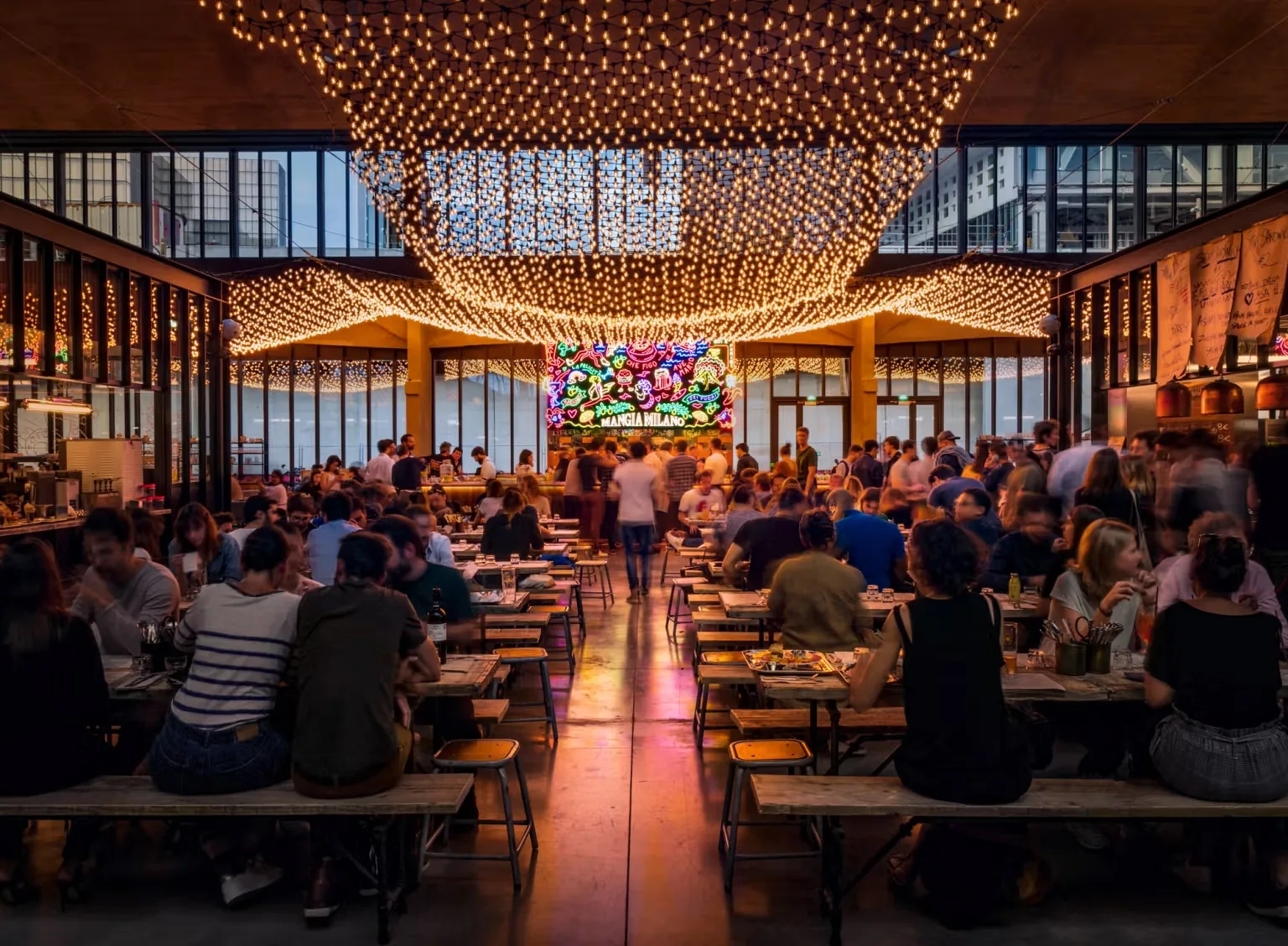 Evening view of long communal dining tables full of guests beneath a canopy of warm string lights and a bright neon sign, at La Felicità, Paris.
