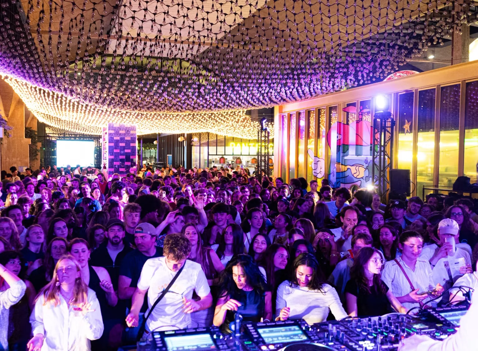 Nighttime crowd dancing under glowing string lights beside a DJ booth during a party, at La Felicità, Paris.