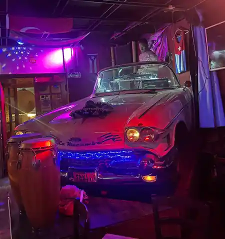 DJ booth behind a vintage car in the neon-lit interior of La Macumba Music Latino Bar, Cuban flags on the wall — salsa, bachata, kizomba, zouk venue in Prague