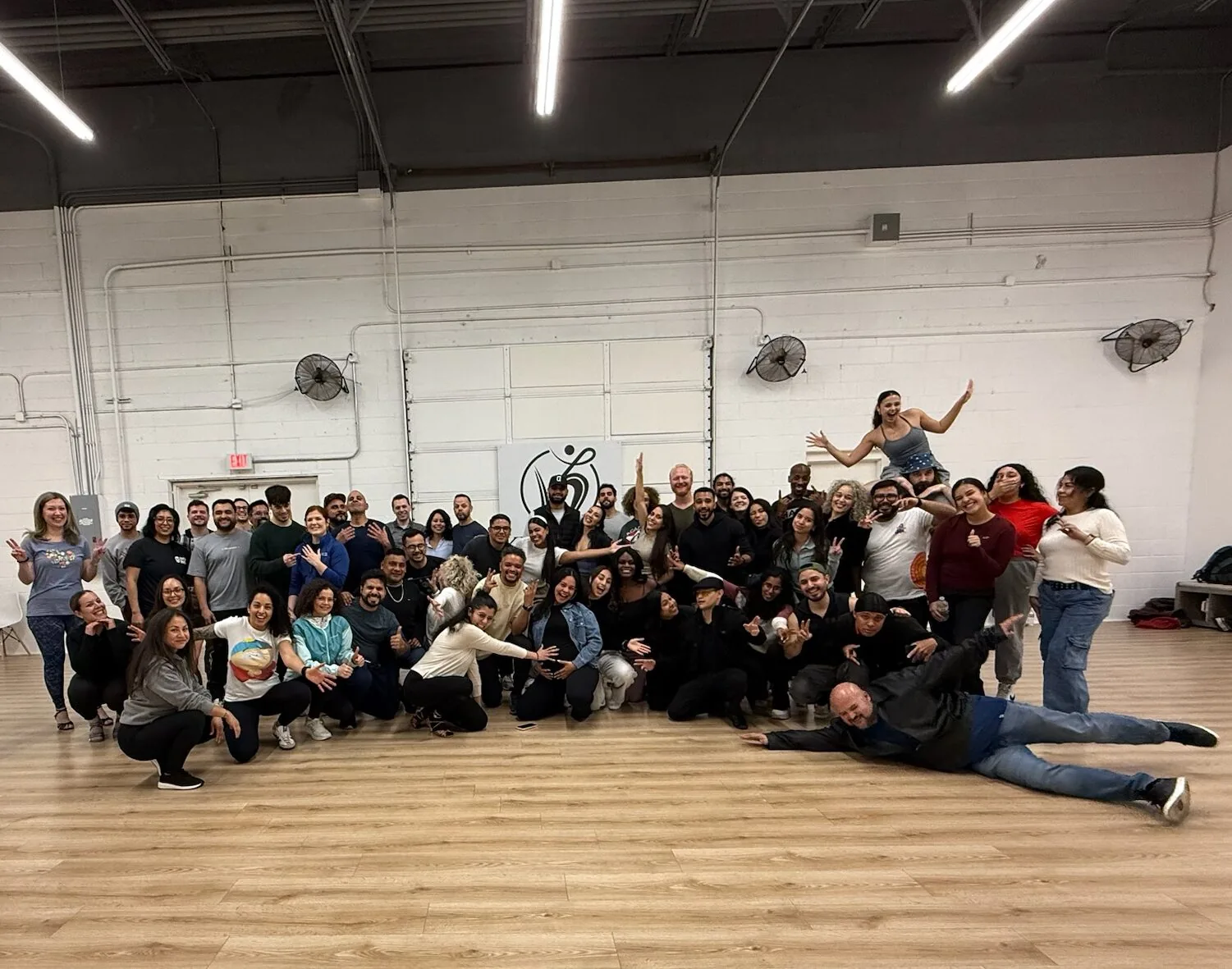 A large group of dancers posing together on the wood floor of the main studio room with white brick walls, wall fans and the Kumbala logo sign, at Kumbala Dance Studio, Farmers Branch.