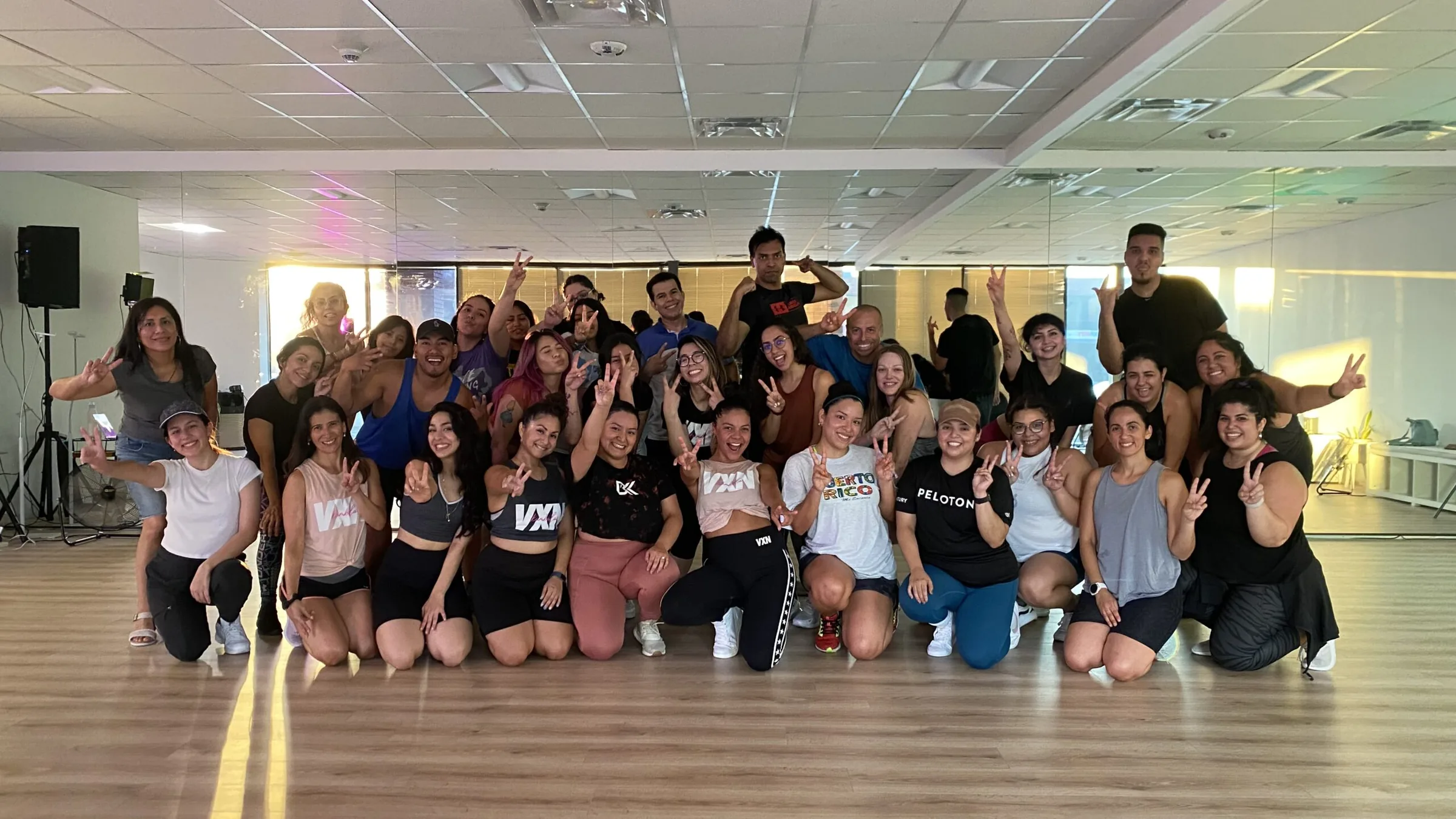 A large group of dancers posing together in a mirrored studio room with wood floors after a class, at Kumbala Dance Studio, Farmers Branch.