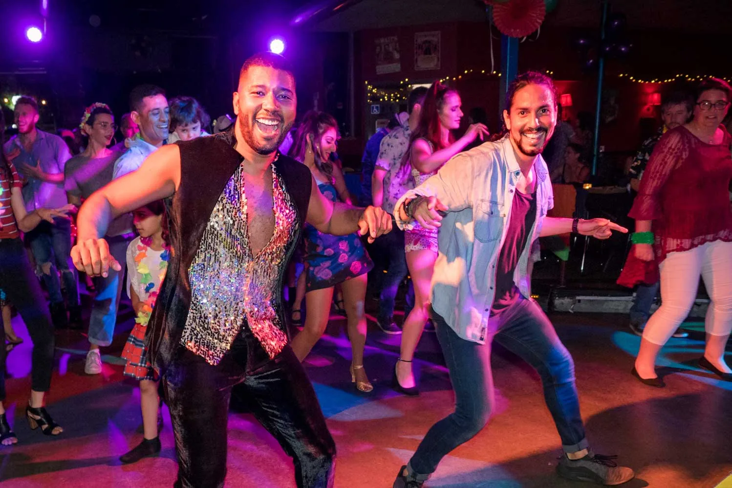 Two men dancing salsa with big smiles on a colorful, lit social dance floor at Latino Grooves Dance Studio in Adelaide