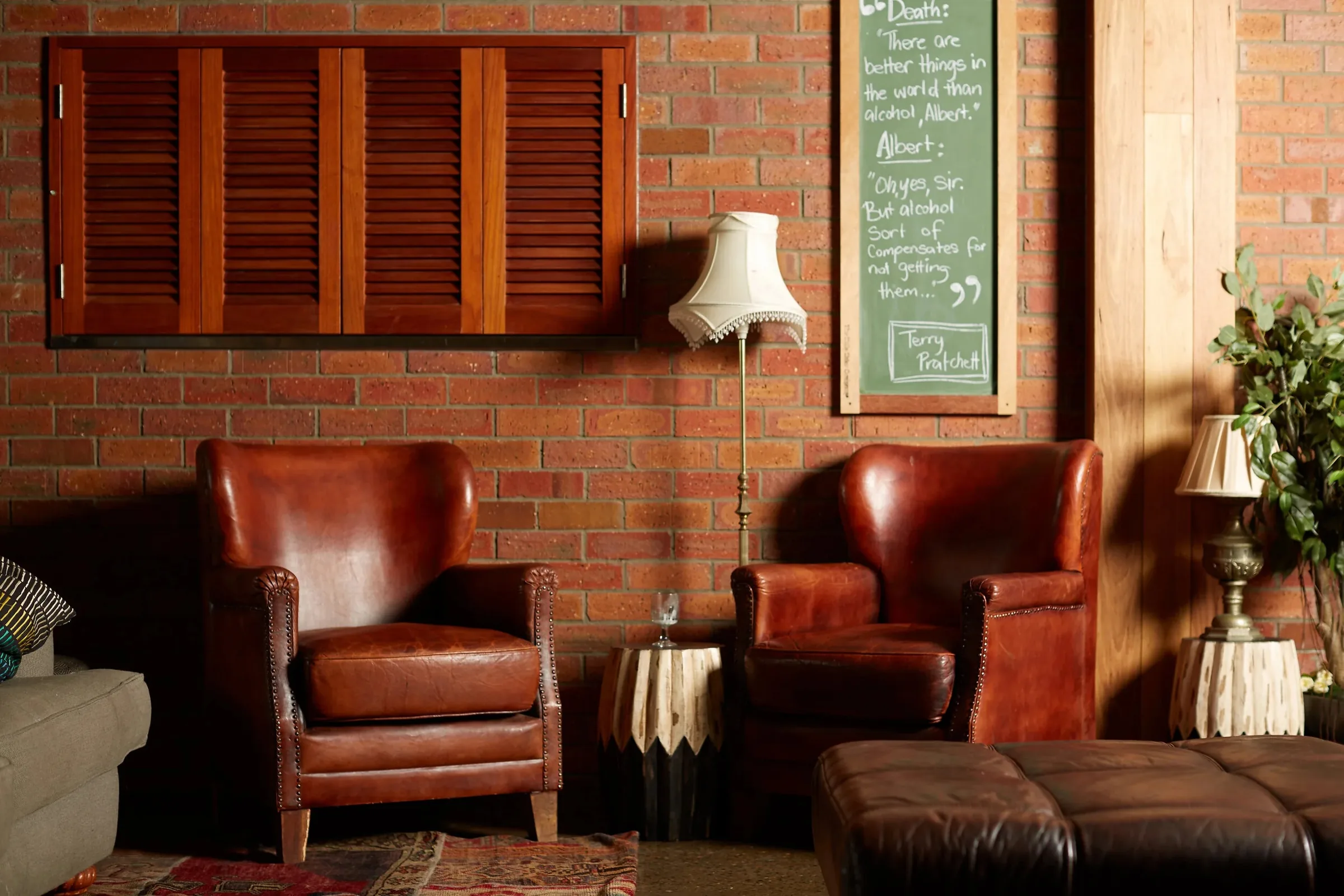 Lounge corner with two vintage tan leather armchairs against an exposed brick wall, a floor lamp and a chalkboard quote at Loft West End, Brisbane.