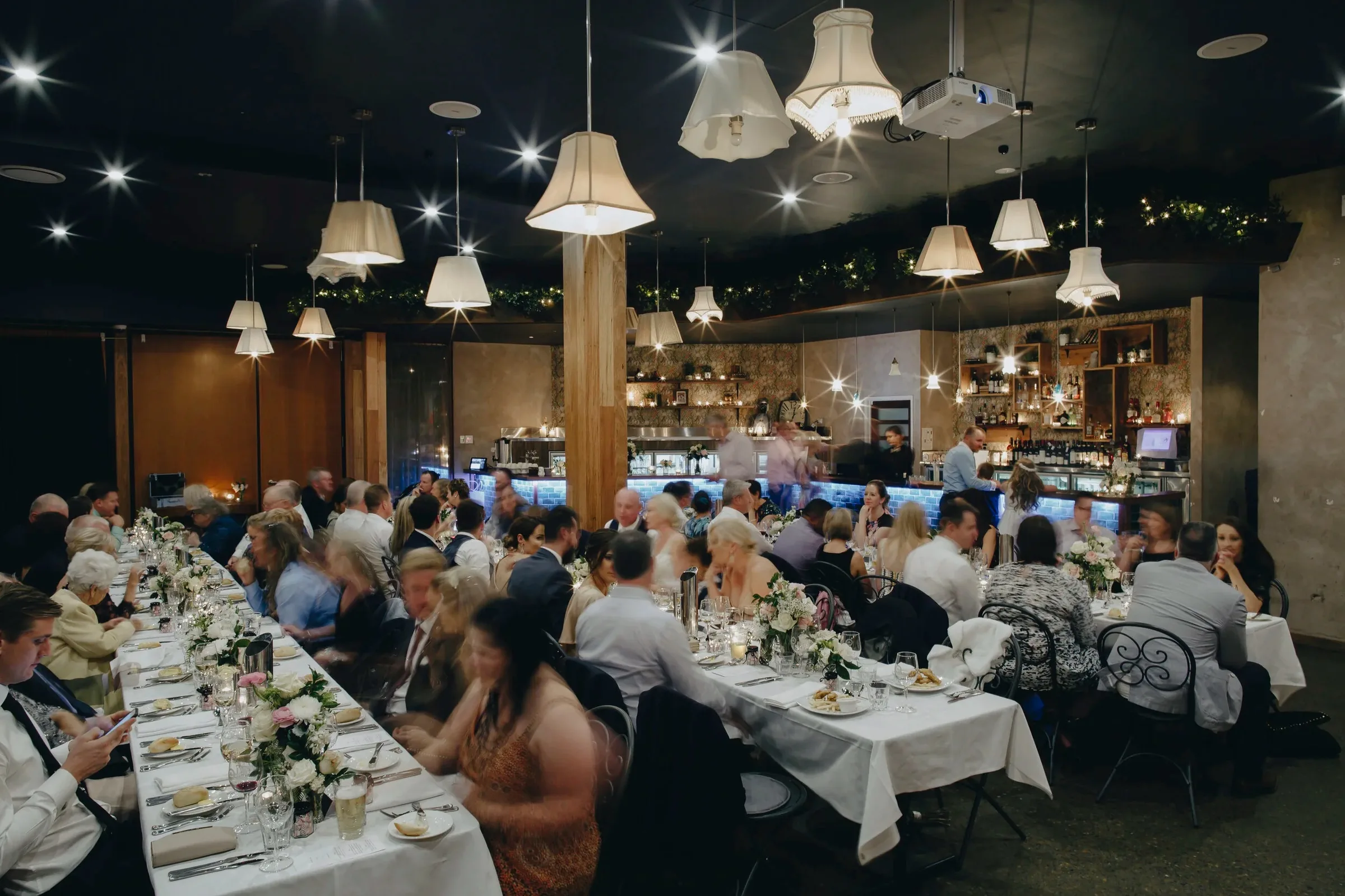 Wide view of the event space set for a seated function with long white-clothed tables, guests dining under pendant lamps and the bar in the background at Loft West End, Brisbane.