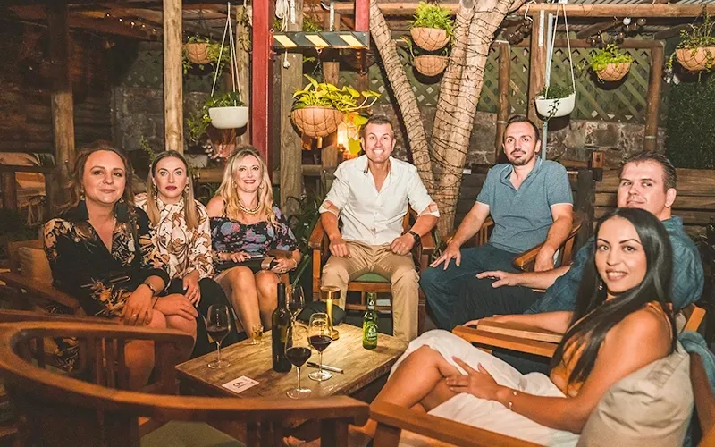 Group of guests seated on timber lounge furniture among hanging baskets and greenery in the covered garden courtyard at The Fourth Monkey Cocktail Bar & Grill, Brisbane.