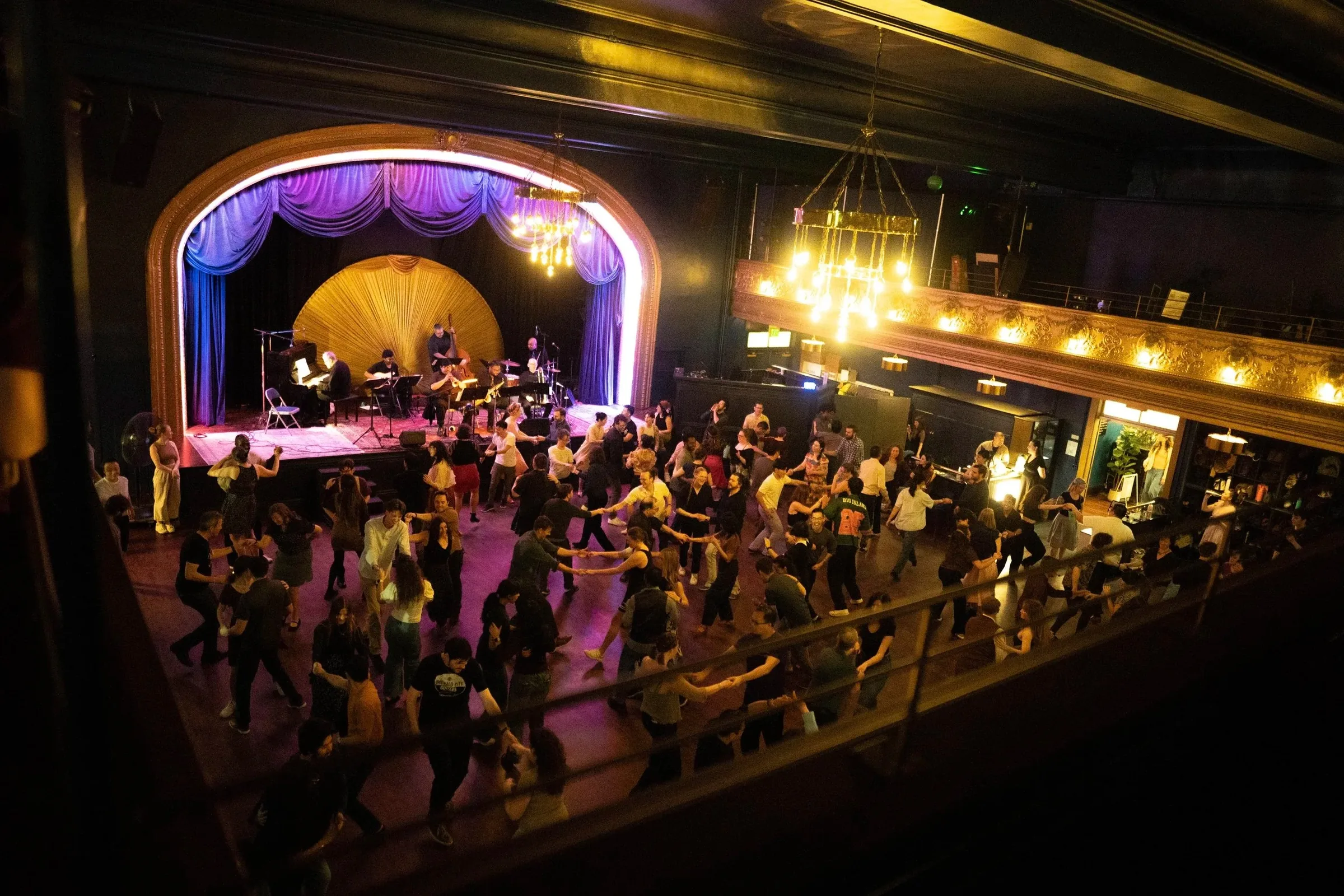 Elevated view of the ballroom during a swing dance night, with dozens of dancers on the wood floor and a live band performing on the arched stage under a glowing chandelier, at Reverie Ballroom, Seattle.