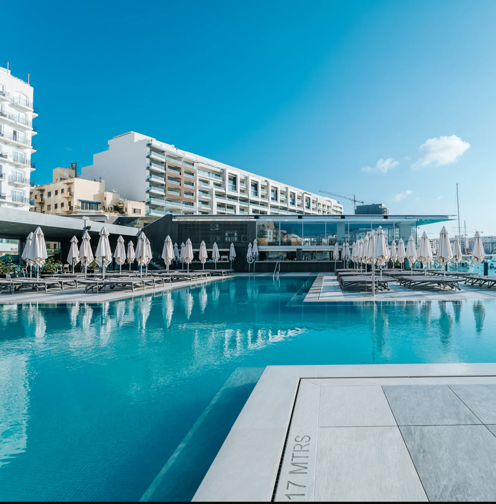 Wide view of the outdoor infinity pool with sun loungers, parasols and the poolside bar overlooking the marina at Aqualuna Beach Club, Malta