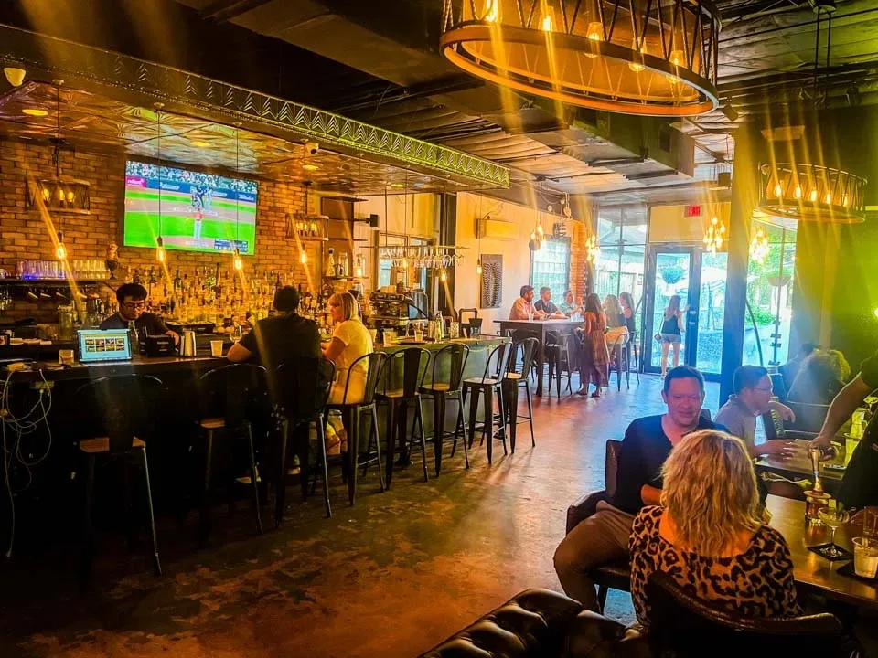 Wide interior view of the bar and lounge with exposed brick walls, industrial pendant lighting, bar-stool seating and seated guests, leading to a glass patio door, at Simone on Sunset in Houston.