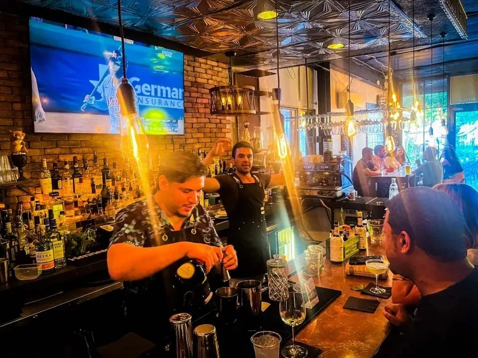 Bartenders in aprons mixing cocktails behind the bar backed by a lit liquor-bottle display and exposed brick, with a sports screen overhead, at Simone on Sunset in Houston.