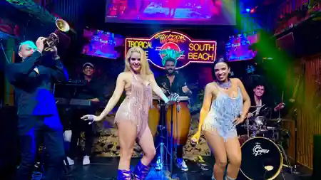 Two showgirls dancing on stage with the live band under the neon Mango's Tropical Cafe South Beach sign in Miami — salsa venue in Miami
