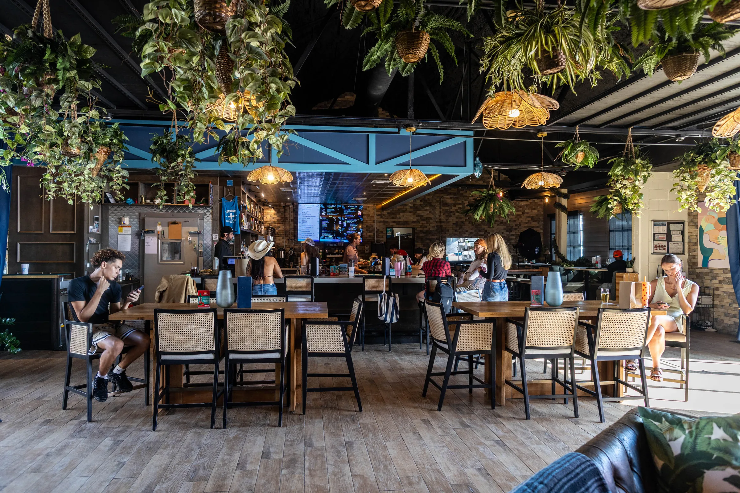 Interior bar and dining hall with long wooden communal tables, cane-back chairs, a full bar, menu screens, and lush hanging plants under the ceiling at Cabana Club, Austin.