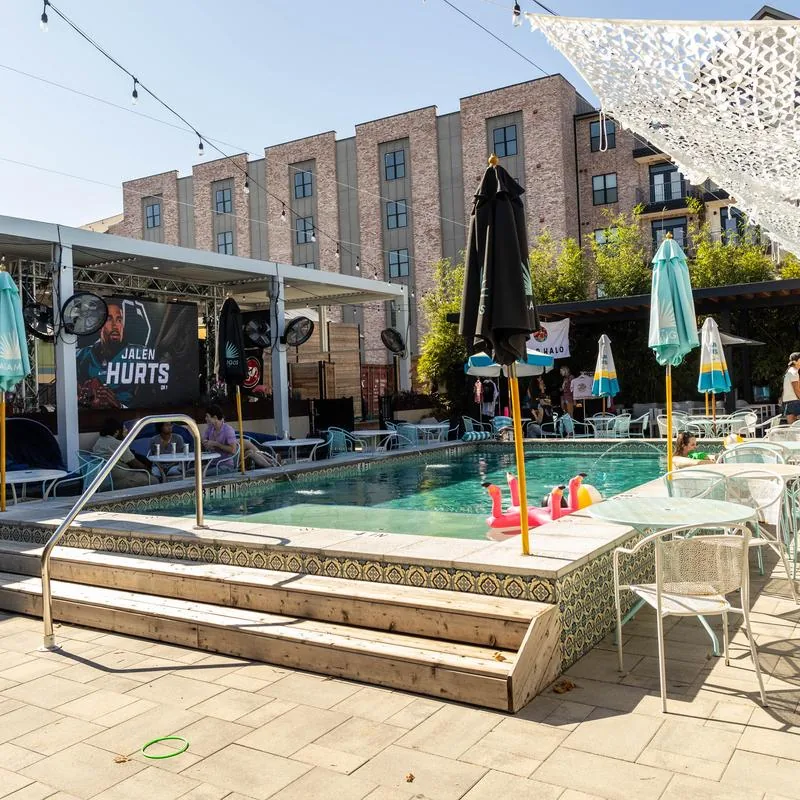 Outdoor pool deck with a small in-ground pool, flamingo floats, teal patio umbrellas, and a large event screen in front of a brick building at Cabana Club, Austin.