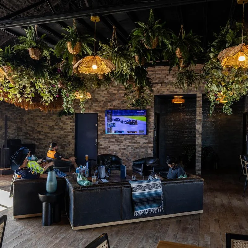 Indoor lounge with black leather sofas, a wall-mounted TV, exposed brick walls, rattan pendant lights, and hanging fern baskets at Cabana Club, Austin.