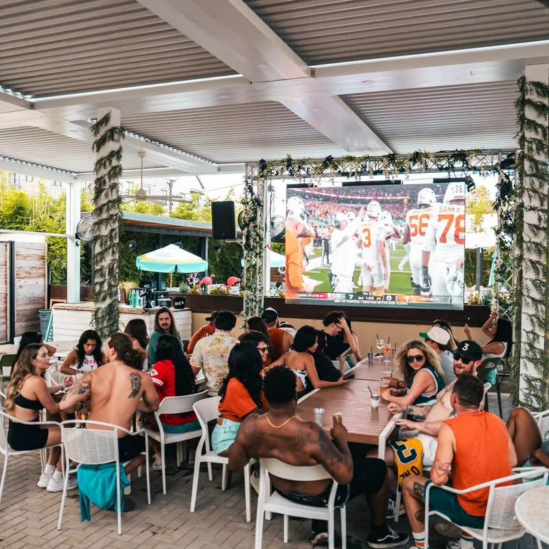 Covered outdoor patio where a crowd watches football on a large screen from a long communal table on a sunny day at Cabana Club, Austin.