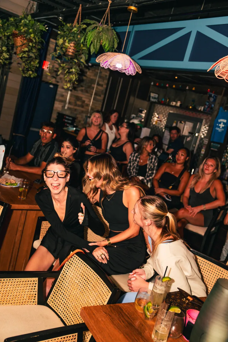 Guests laughing over cocktails in the plant-filled indoor lounge in the evening at Cabana Club, Austin.