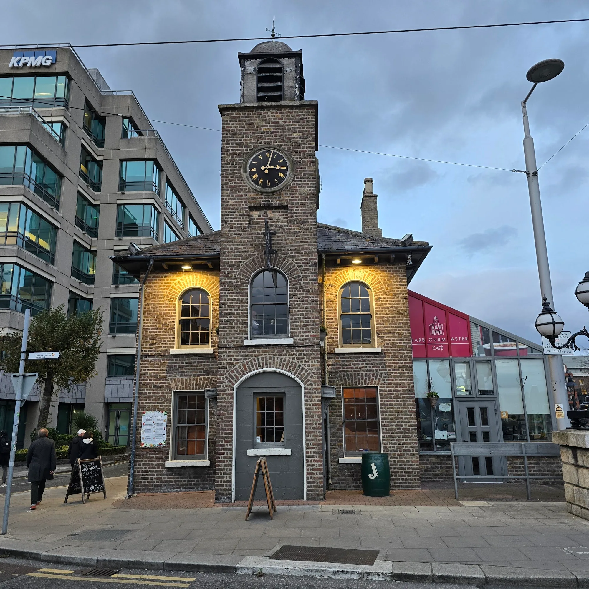 Dusk exterior of the Harbourmaster clock tower building in Dublin, softly lit with red cafe signage and people passing on the dockland street