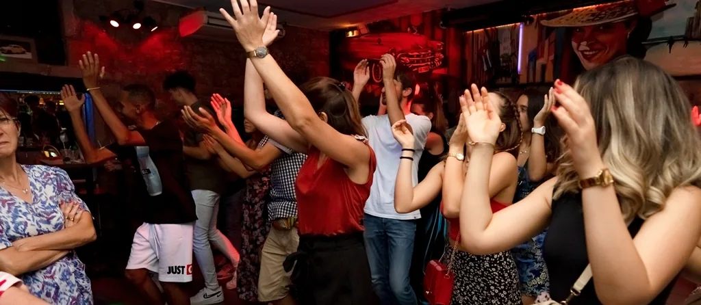Crowd dancing with hands raised under red lighting, surrounded by Cuban-themed murals and decor at Cabana Cafe, Lyon.