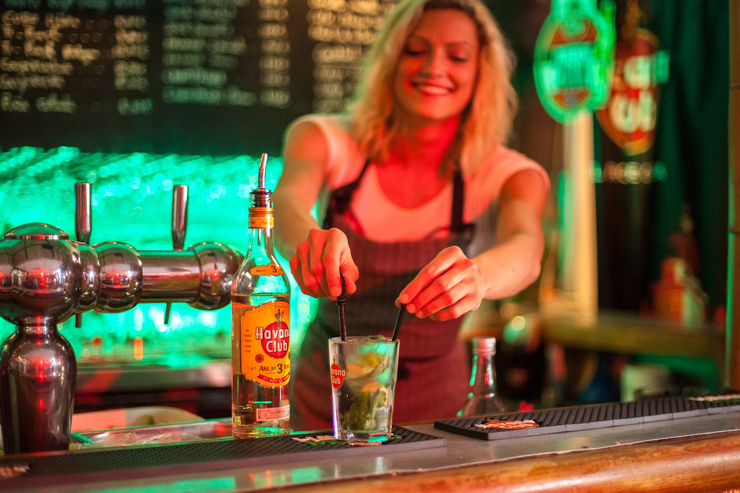 Bartender preparing a mojito behind the bar with beer taps and a bottle of Havana Club rum under green neon light at Cabana Cafe, Lyon.