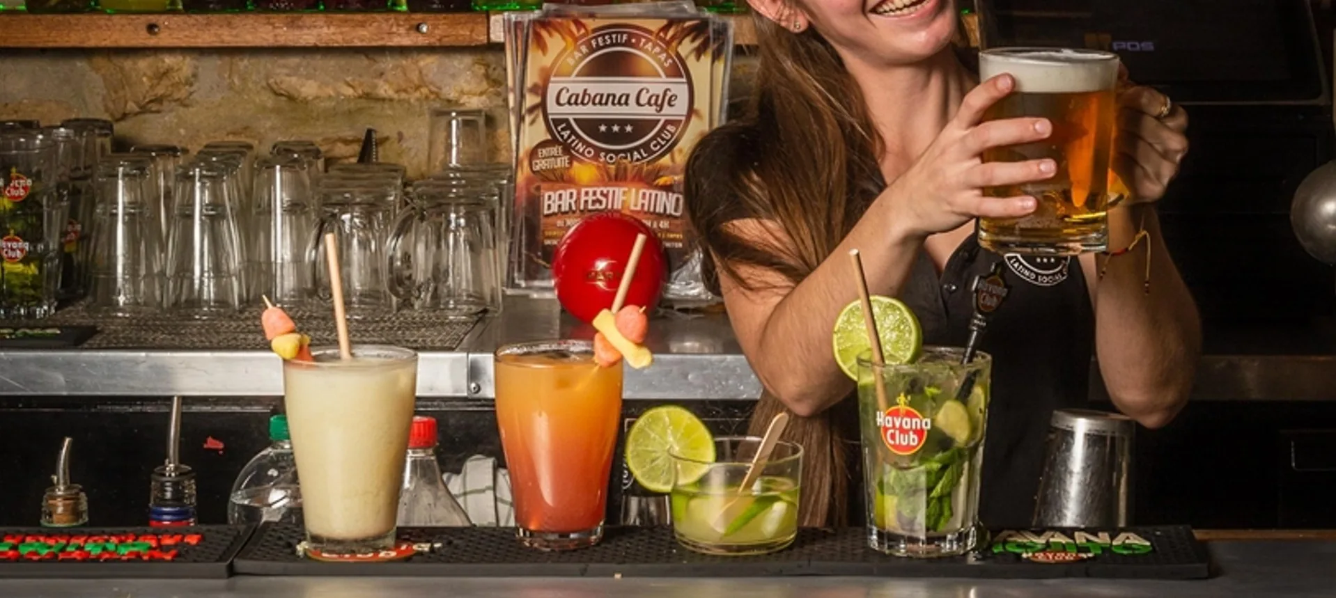 Bartender serving a beer at the bar counter lined with cocktails and mojitos beside a Cabana Cafe latino social club card at Cabana Cafe, Lyon.