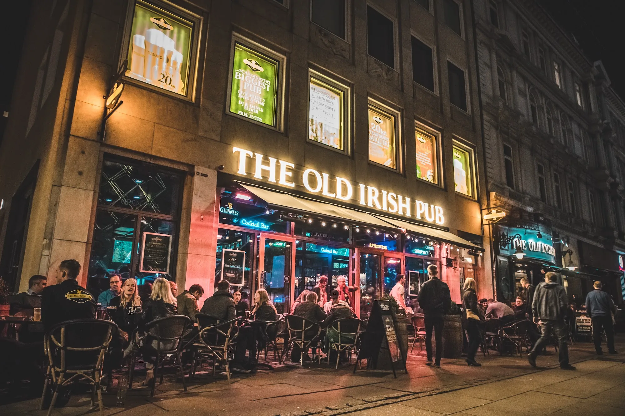 The Old Irish Pub Copenhagen exterior lit up at night with illuminated signage and a busy crowd at outdoor tables, salsa, bachata venue in Copenhagen
