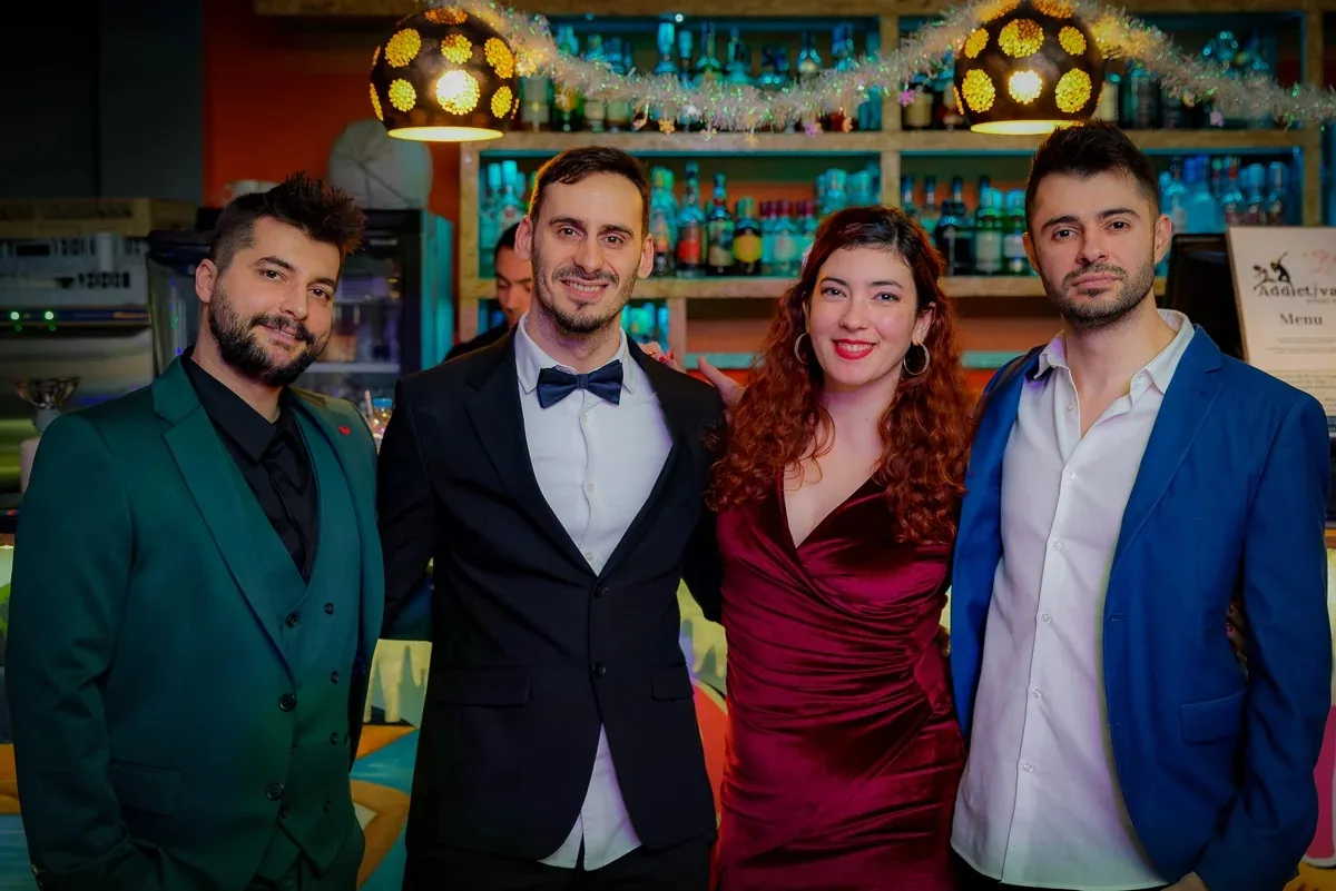Four people in formal evening wear posing together in front of the venue's colorful backlit bottle shelves and disco-ball pendant lamps, Addictiva Athens Bar, Athens