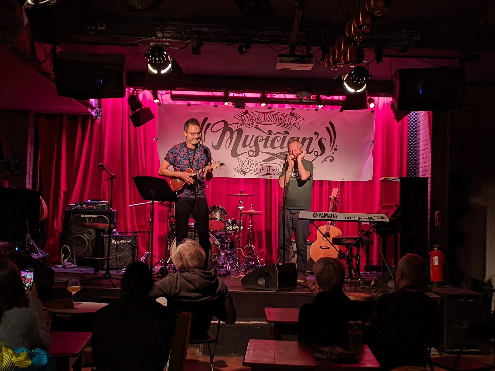 Live band performing on the pink-lit stage under a Brussels Musicians Meet-Up banner at The Embassy Room Brussels with a seated audience