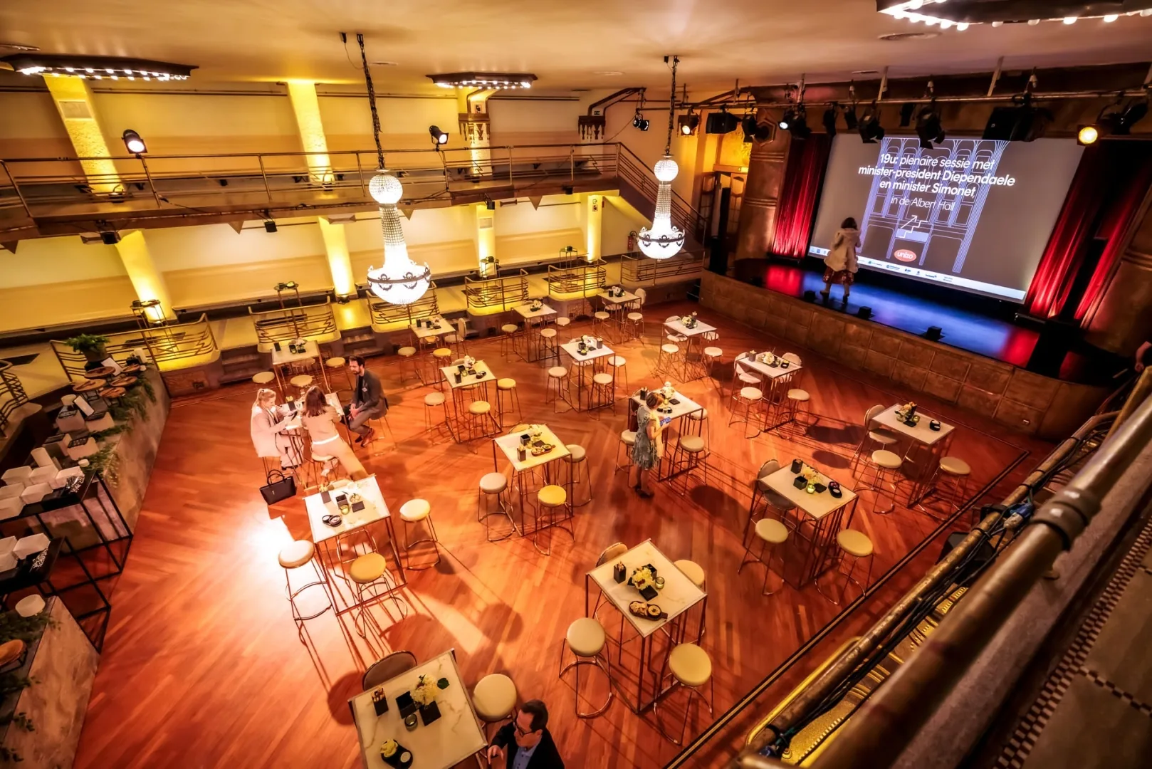 Grand Albert Hall Brussels main hall from above showing the wooden floor, cocktail tables, chandeliers and a stage with a large screen