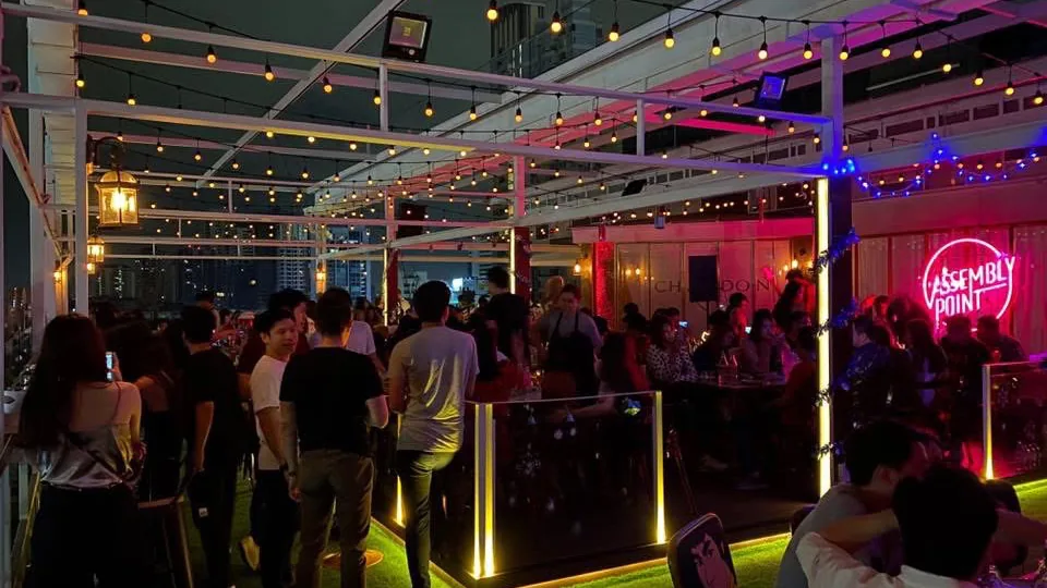 Rooftop bar crowd under Assembly Point neon sign at night, Bangkok